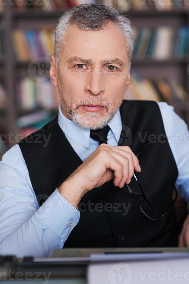 Confident author. Confident grey hair senior man in formalwear sitting at the typewriter and looking at camera with bookshelf in the background photo