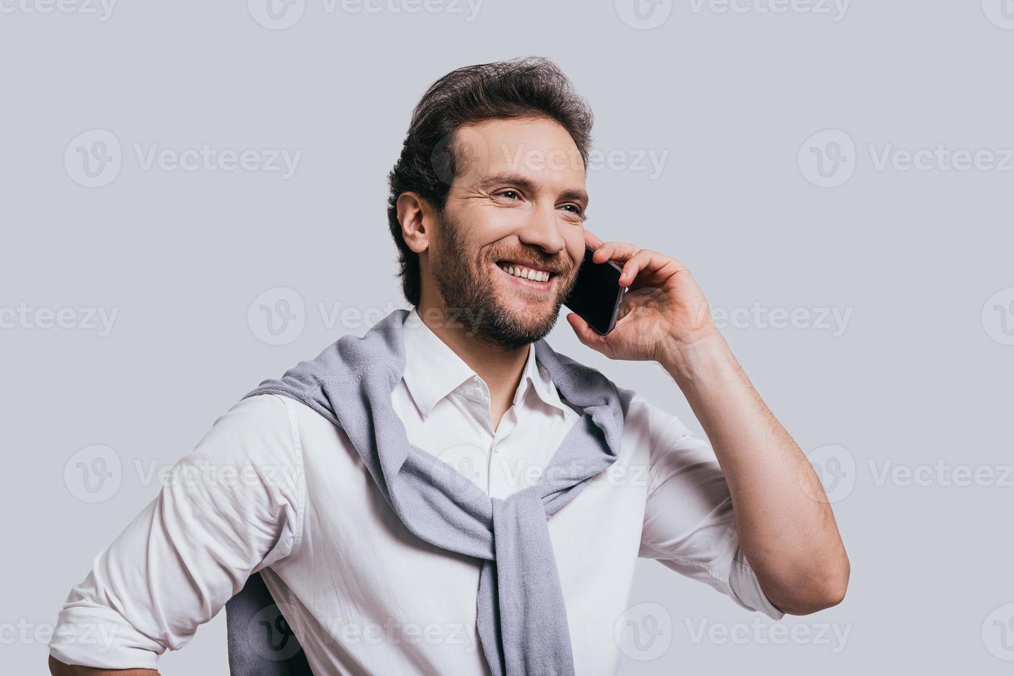 Pleasant business talk. Beautiful young man in smart casual clothes talking on the phone and smiling while standing against grey background photo