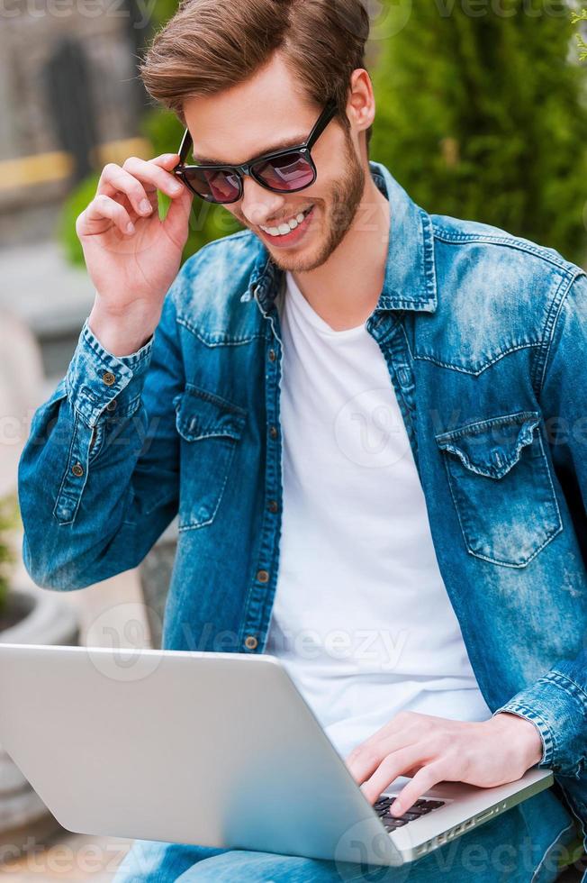 Enjoying free Wi-Fi. Cheerful young man working on laptop and adjusting his eyewear while sitting outdoors photo