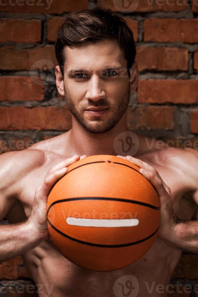 He is willing to this game. Handsome young muscular man holding basketball ball while standing against brick wall photo