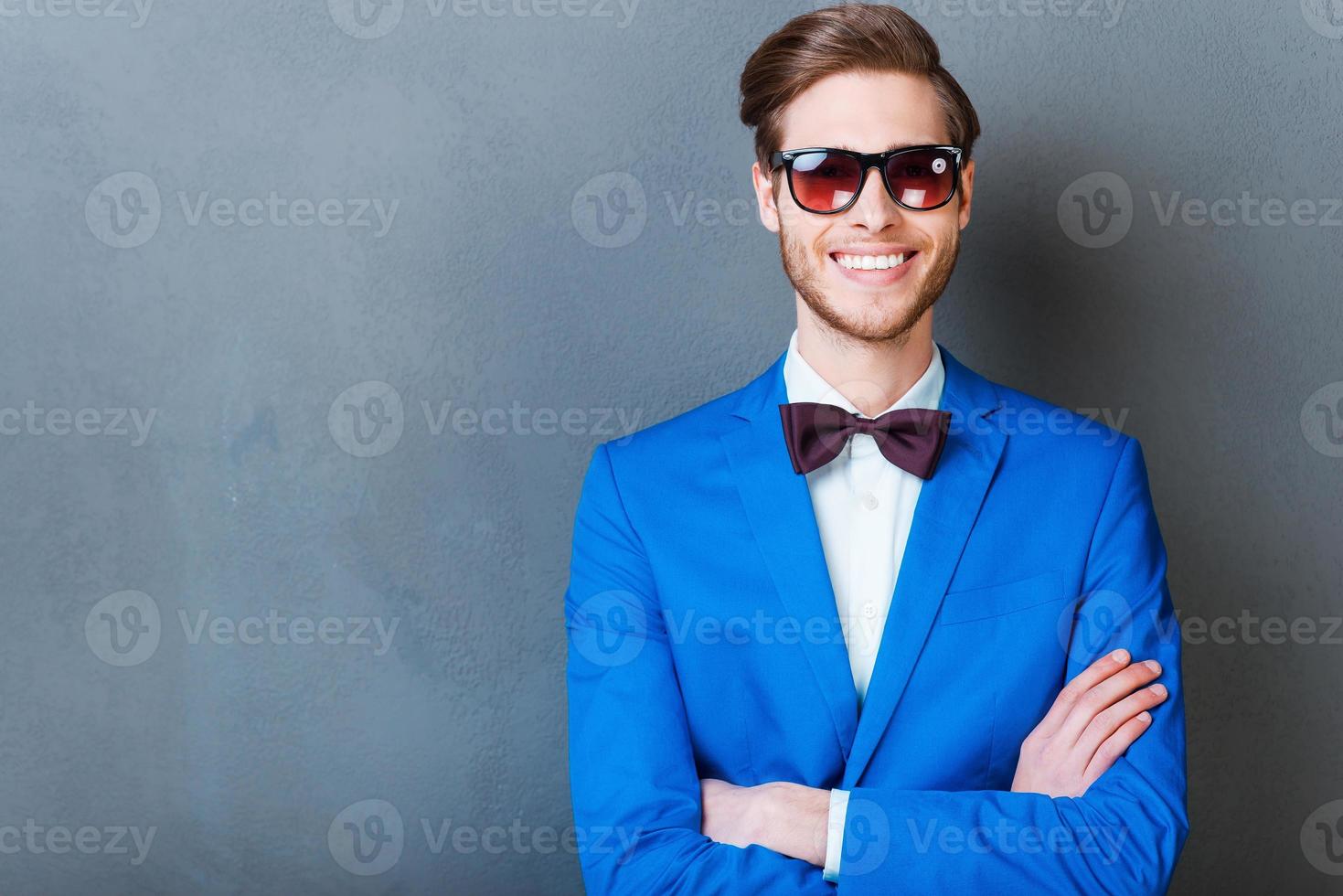 Smile is a part of his style. Cheerful young man in eyewear keeping arms crossed and smiling at camera while standing against grey background photo