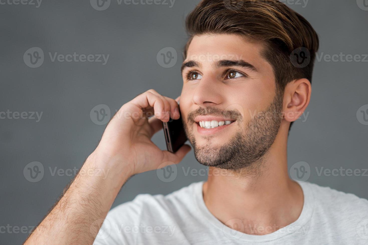 Good talk with friend. Happy young man talking on mobile phone and smiling while standing against grey background photo