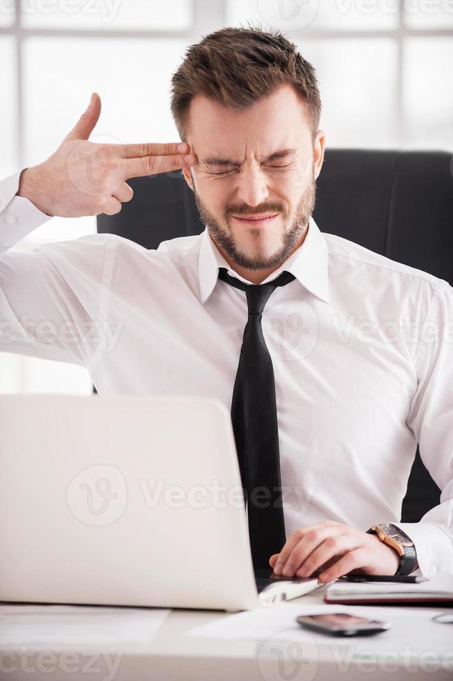 Monday morning suicide. Frustrated young man in formalwear touching his temple with finger gun and keeping eyes closed while sitting at his working place photo