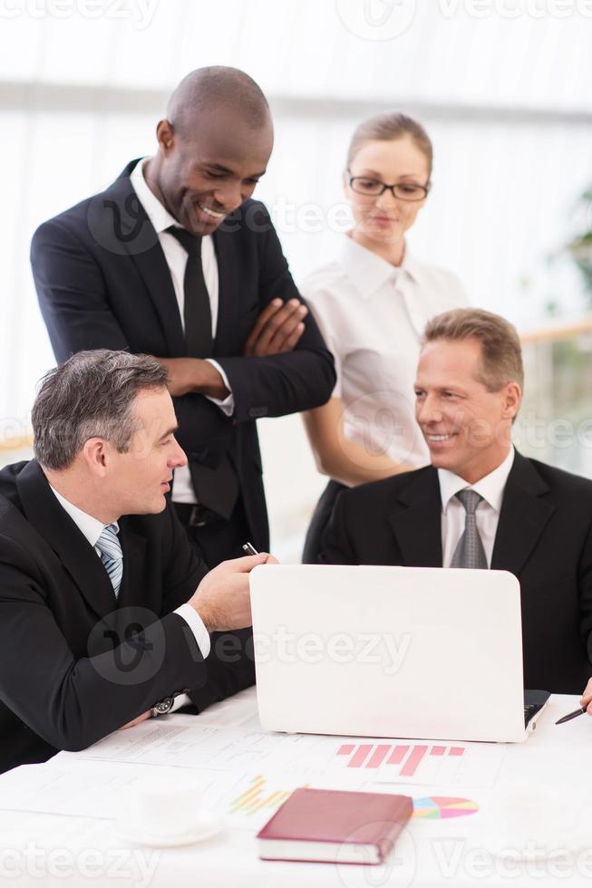 Business team at meeting. Two cheerful mature man in formalwear sitting at the table and communicating while two another people standing close to them photo