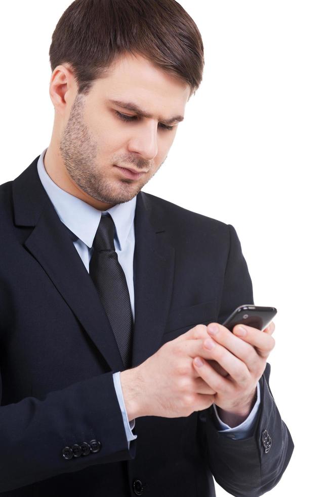 Typing a business message. Confident young man in formalwear holding mobile phone and looking at it while standing isolated on white photo