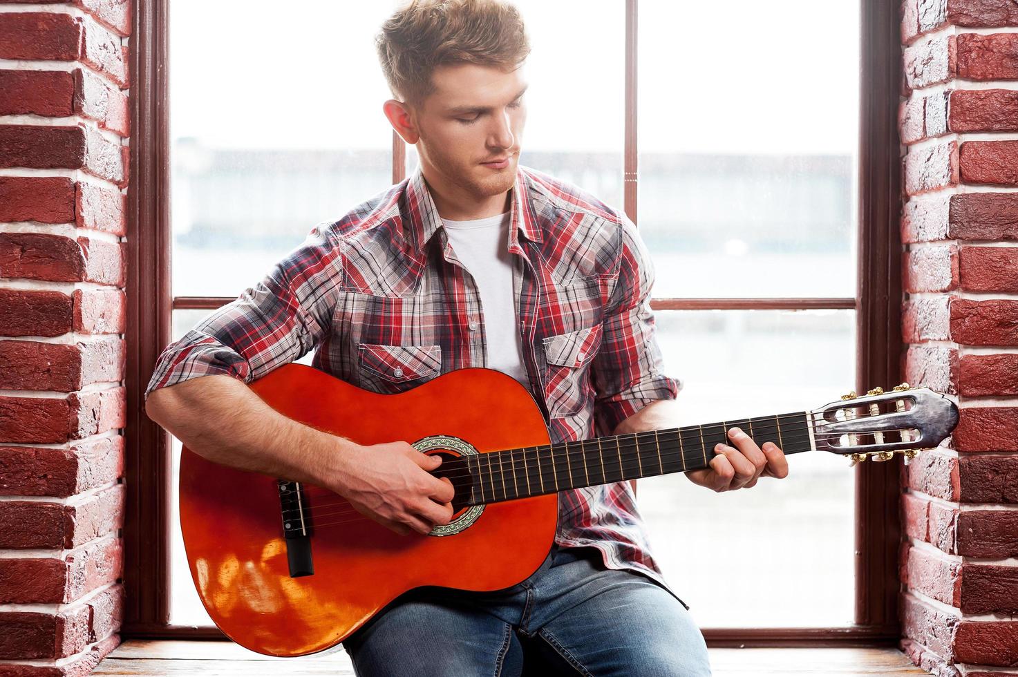 tiene alma creativa. apuesto joven tocando la guitarra acústica mientras se sienta en el alféizar de la ventana foto