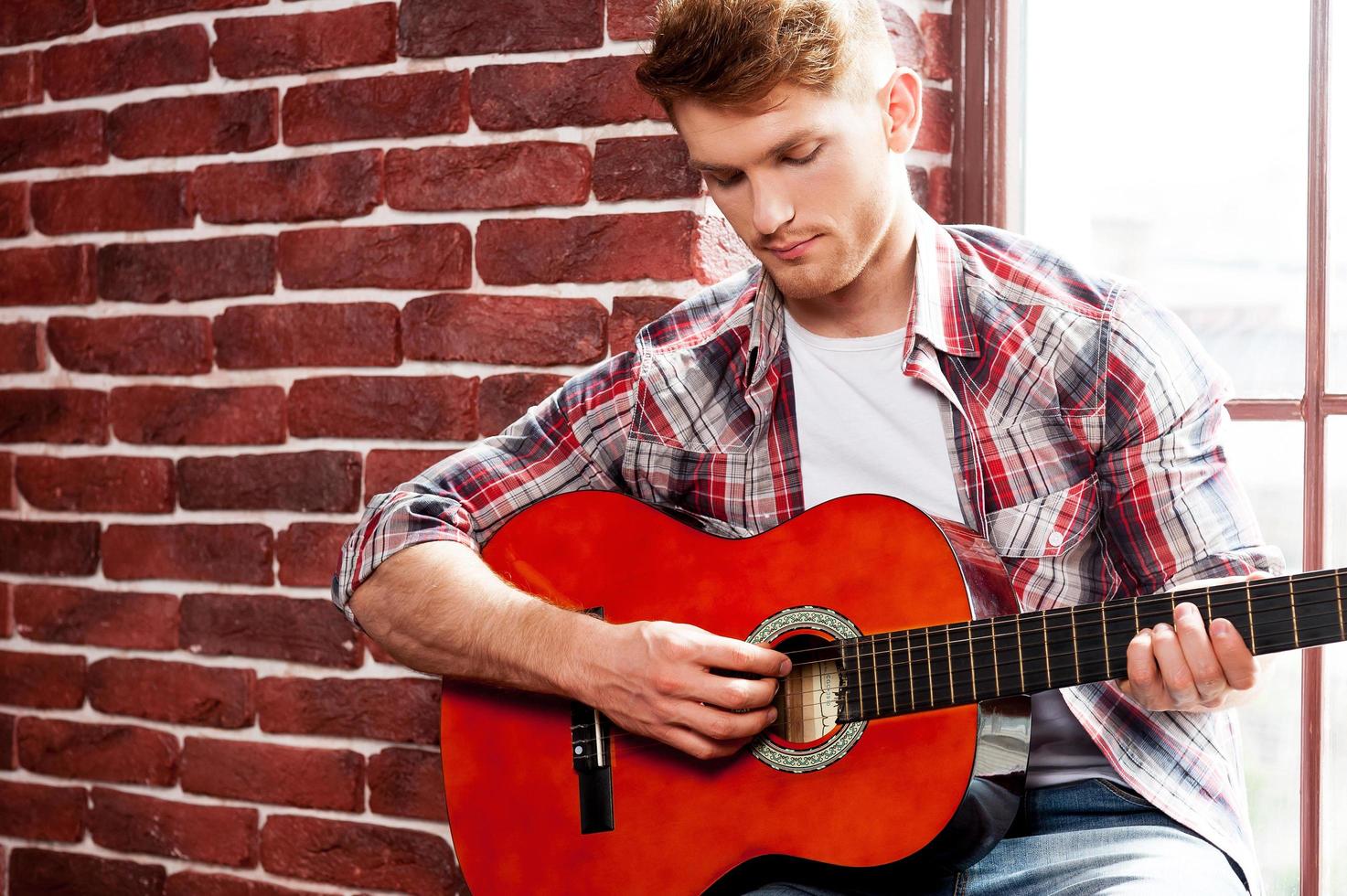 Creative soul. Handsome young man playing acoustic guitar while sitting on the window sill photo