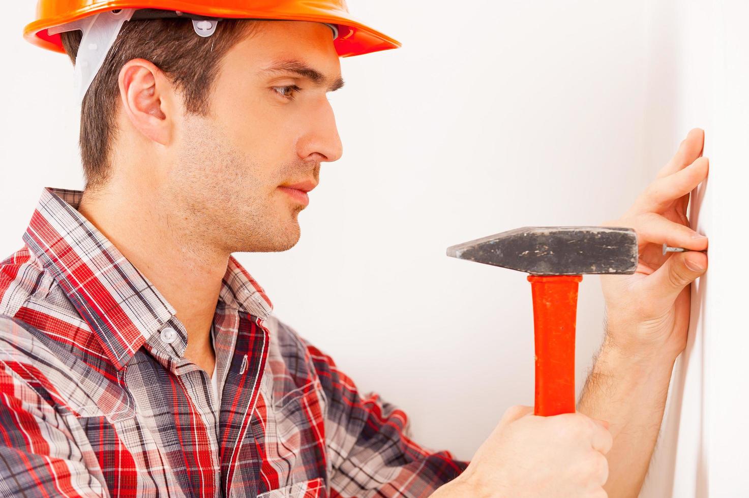 Home improvement. Side view of handsome young handyman in hardhat hammering a nail while standing near the wall photo