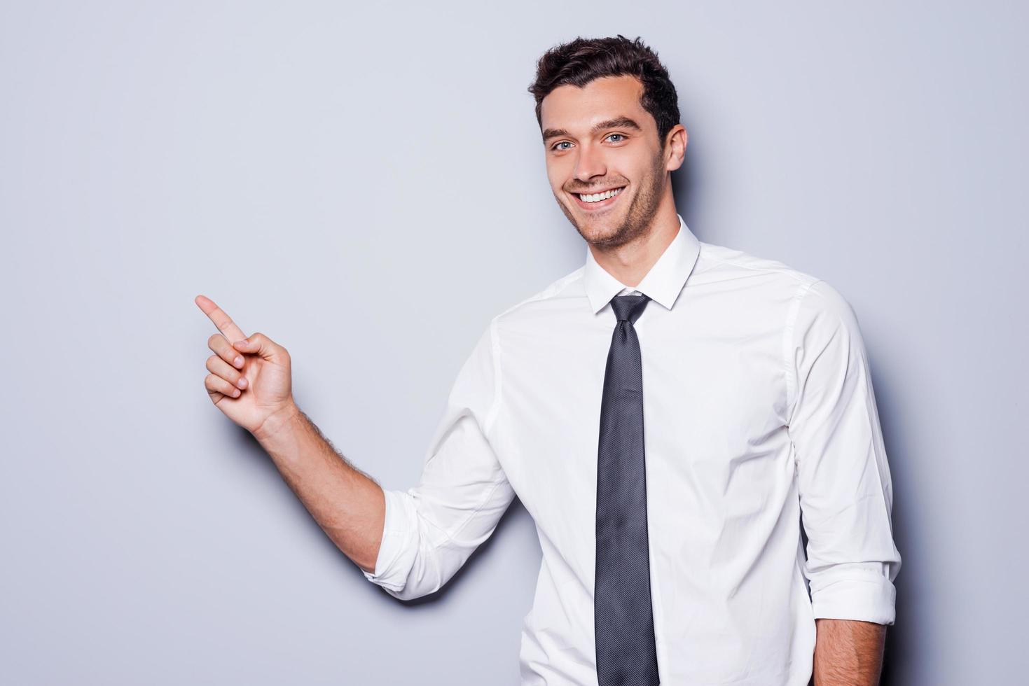 hombre de negocios que señala el espacio de la copia. joven feliz con camisa y corbata mirando a la cámara y sonriendo mientras se enfrenta a un fondo gris y señala el espacio de copia foto