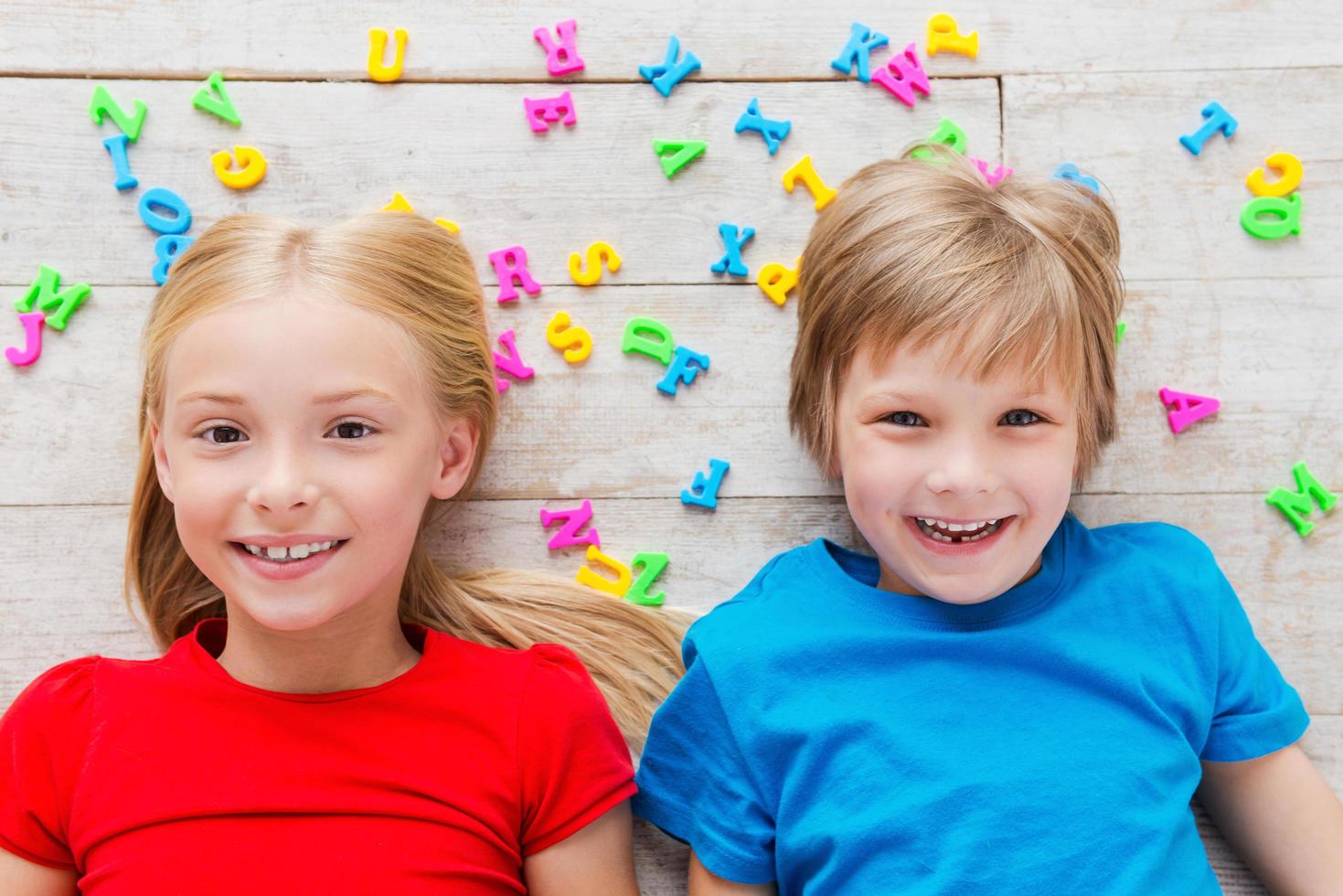 Cheeky kids. Top view of two cute little children looking at camera and smiling while lying on the floor with plastic colorful letters laying around them photo