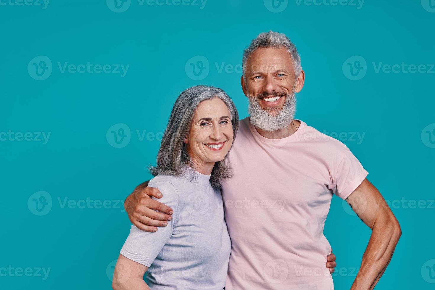 Happy senior couple looking at camera and smiling while standing together against blue background photo