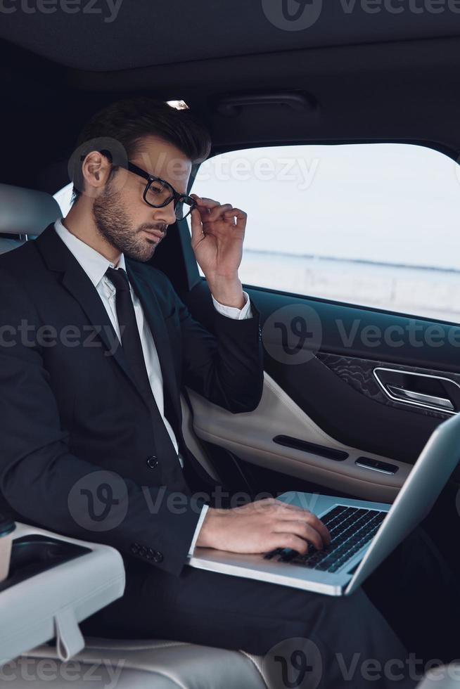 el negocio es su vida. un joven pensativo con traje completo que trabaja con una laptop y se ajusta los anteojos mientras está sentado en el auto foto