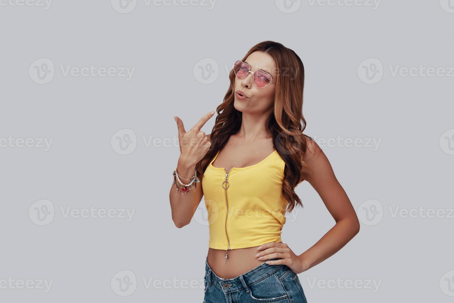 Attractive young woman looking at camera and blowing on hand gun while standing against grey background photo