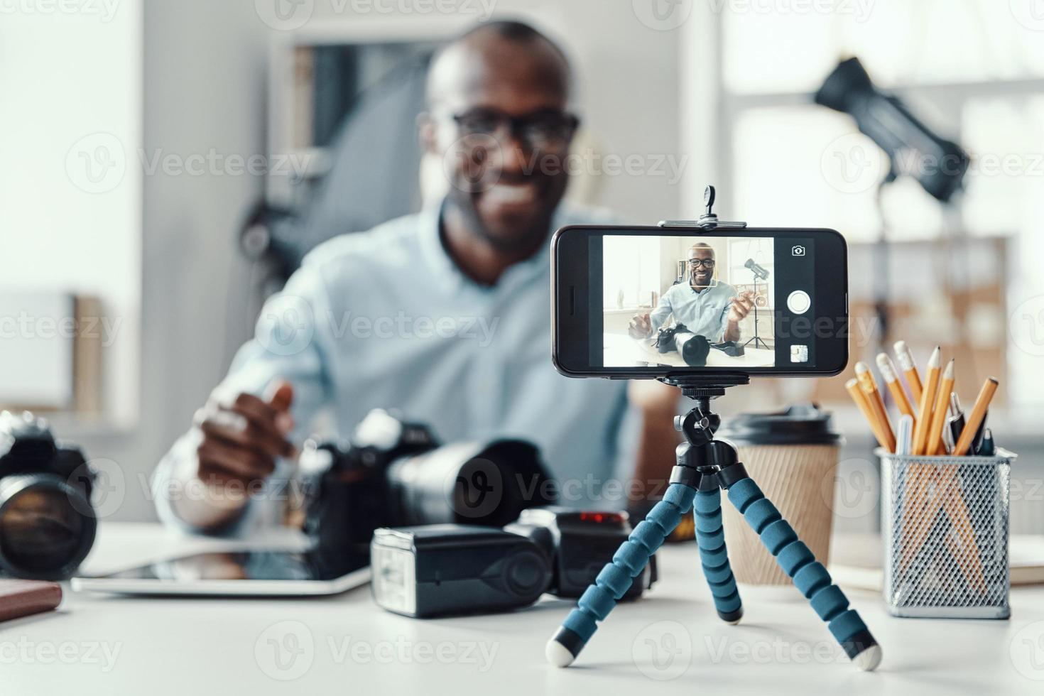 Happy young African man in shirt showing digital camera and telling something while making social media video photo