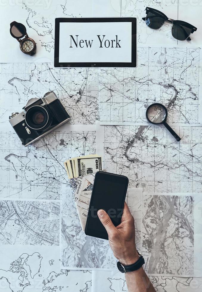 New York is waiting Close up top view of man holding smart phone, passport and money with sunglasses, photo camera, compass, magnifying glass lying on map around