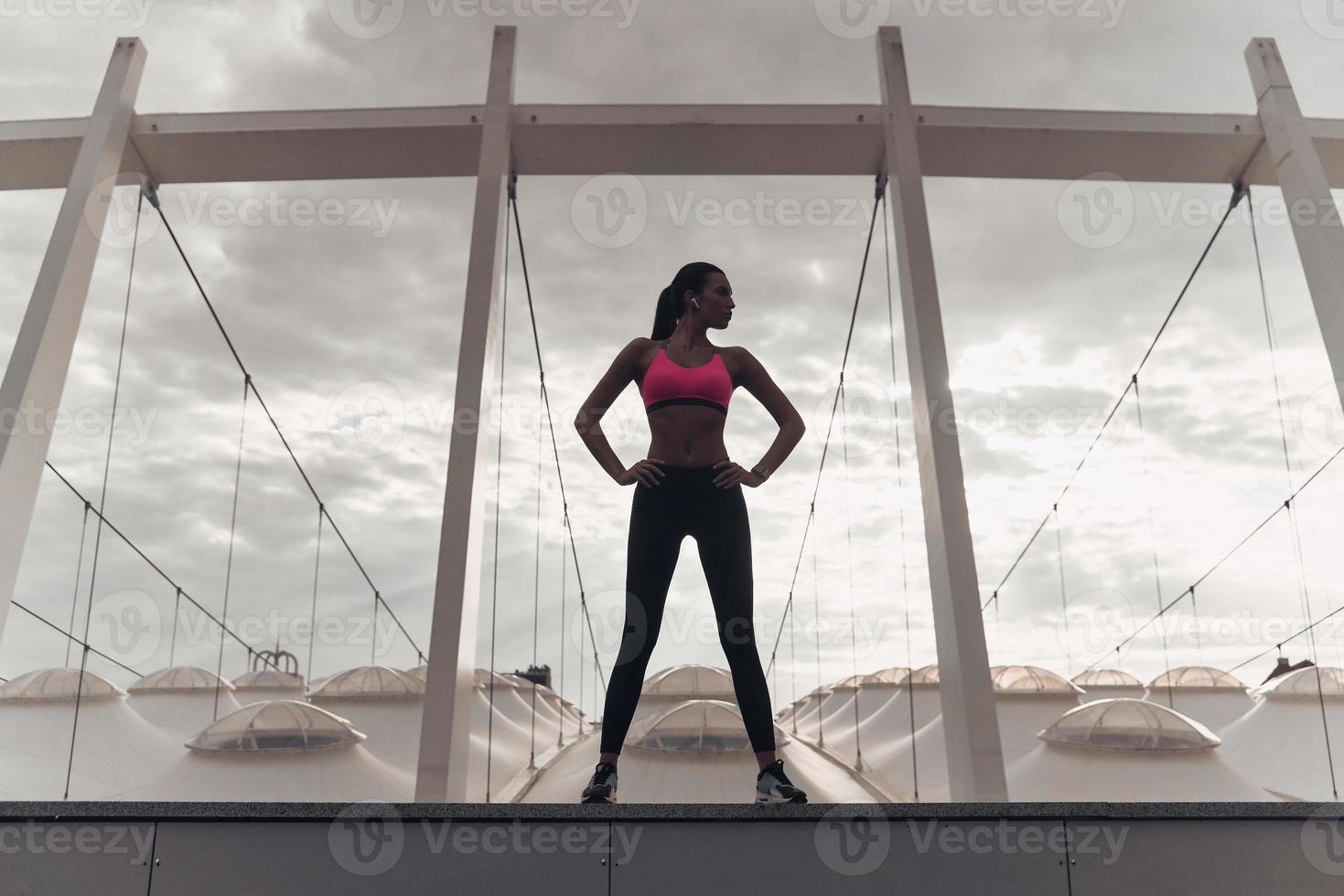 Strong and confident. Full length of modern young woman in sports clothing keeping hands on hips and looking away while standing outdoors photo