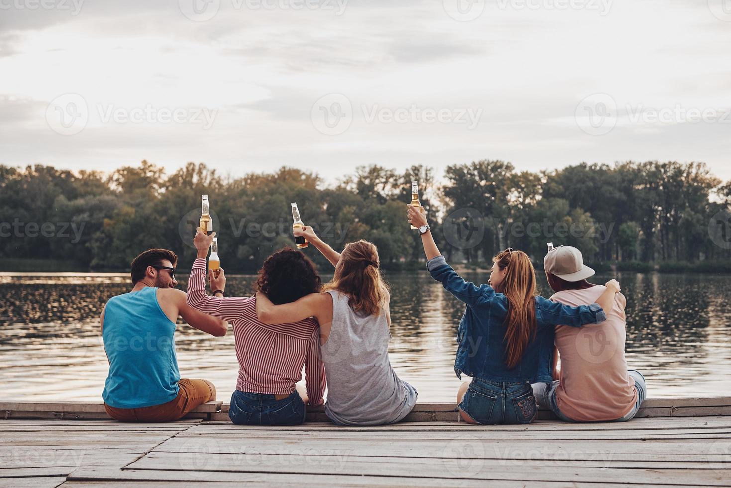 a la interminable vista trasera de verano de jóvenes con ropa informal brindando con botellas de cerveza mientras se sientan en el muelle foto