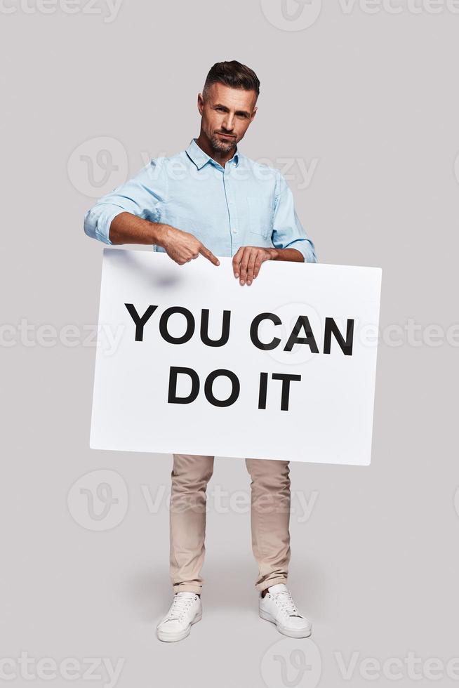 I believe in you Full length of handsome young man holding poster and gesturing while standing against grey background photo
