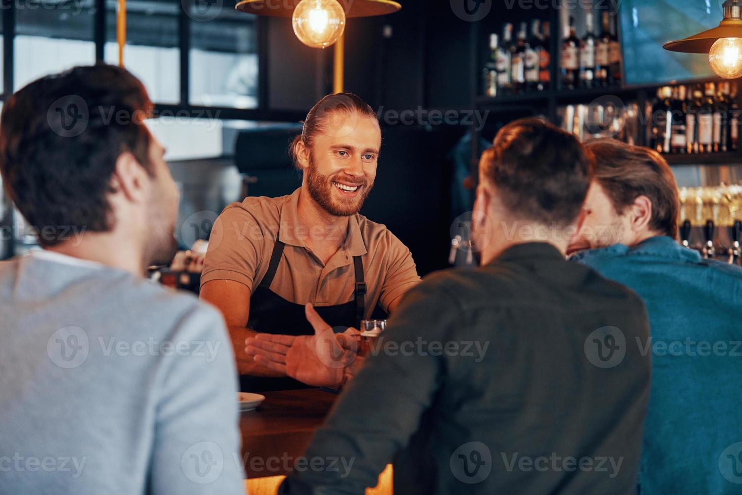 camarero alegre que sirve cerveza a los jóvenes mientras está de pie en el mostrador del bar en el pub foto