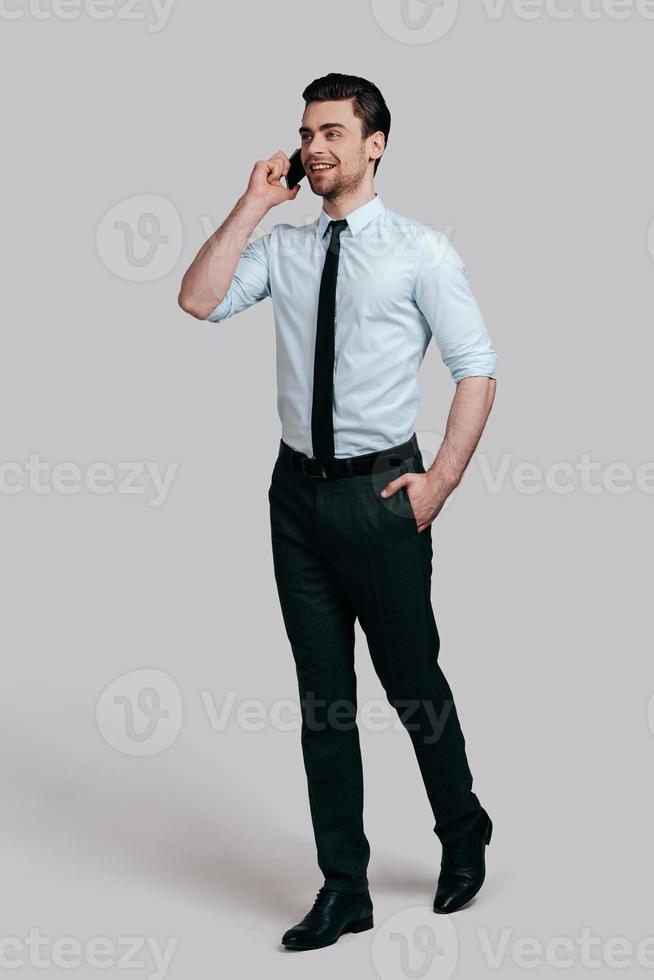 Pleasant business talk. Full length of handsome young man talking on smart phone and keeping hand in pocket while standing against grey background photo