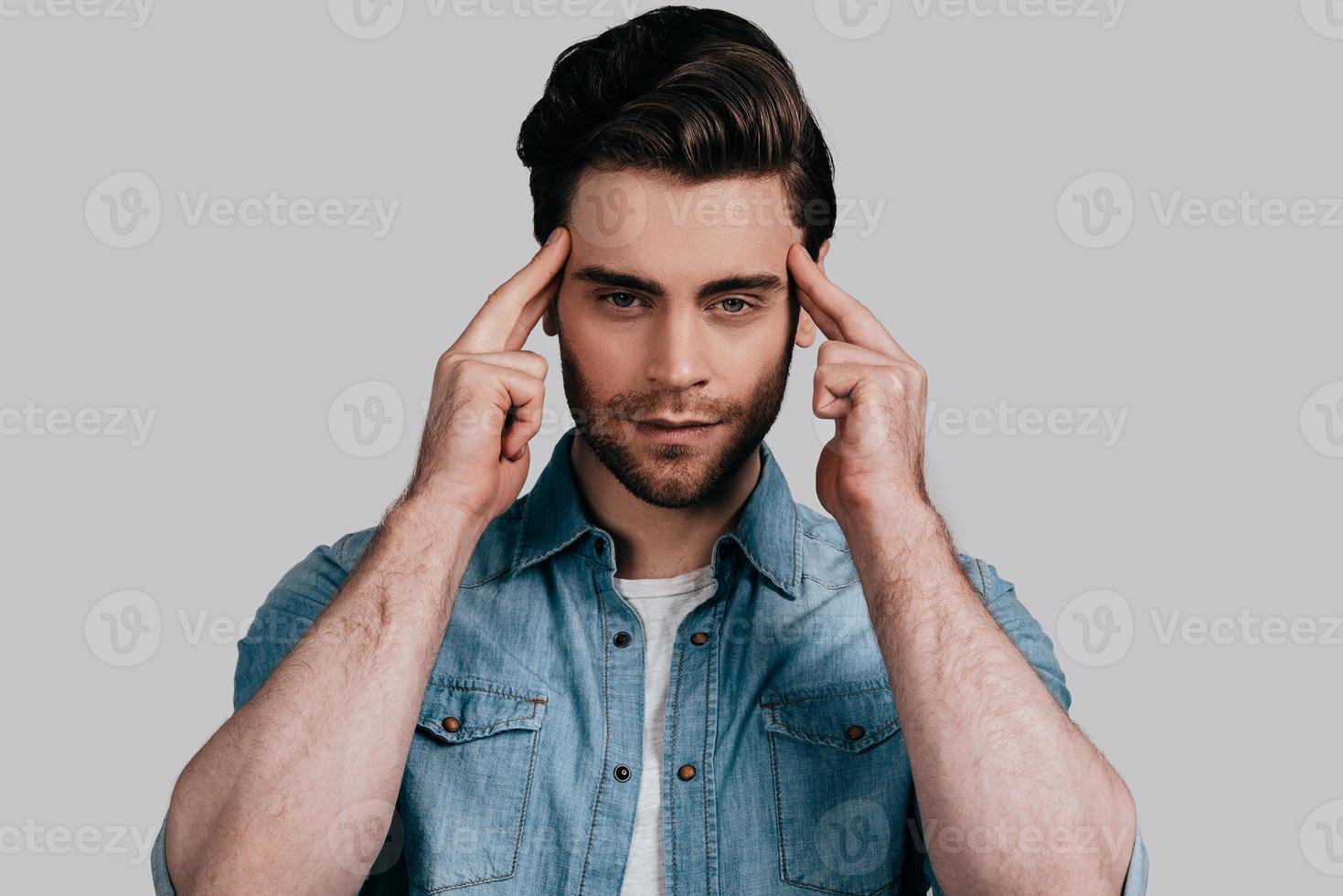 Feeling stressed. Frustrated young man in casual wear holding fingers on head and looking at camera while standing against grey background photo