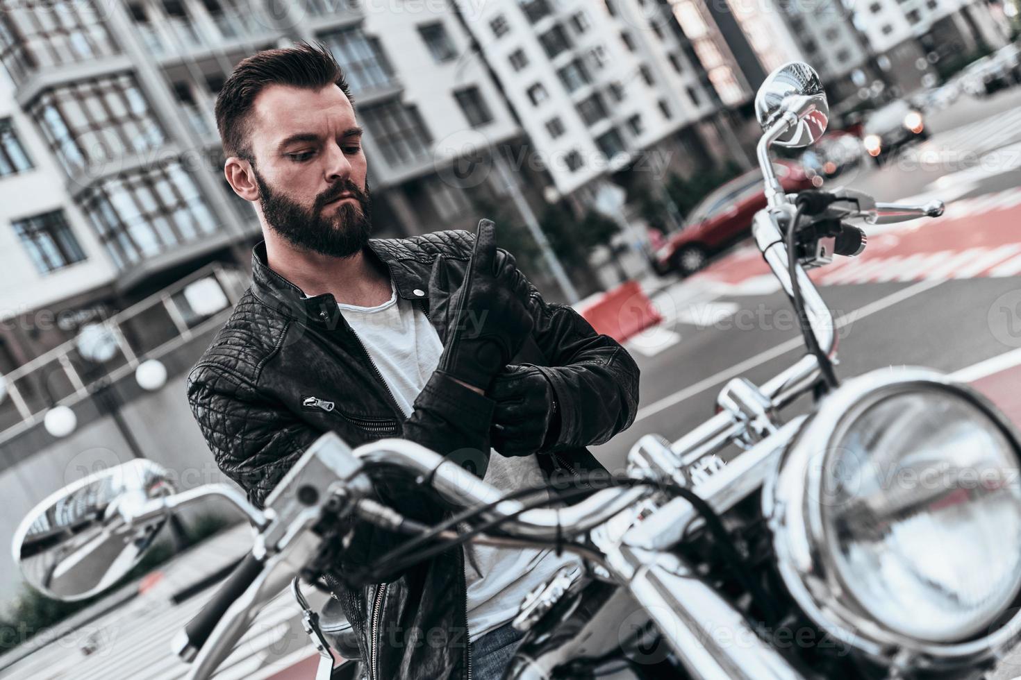 In his own style. Handsome young man taking on protective gloves while sitting on the motorbike outdoors photo