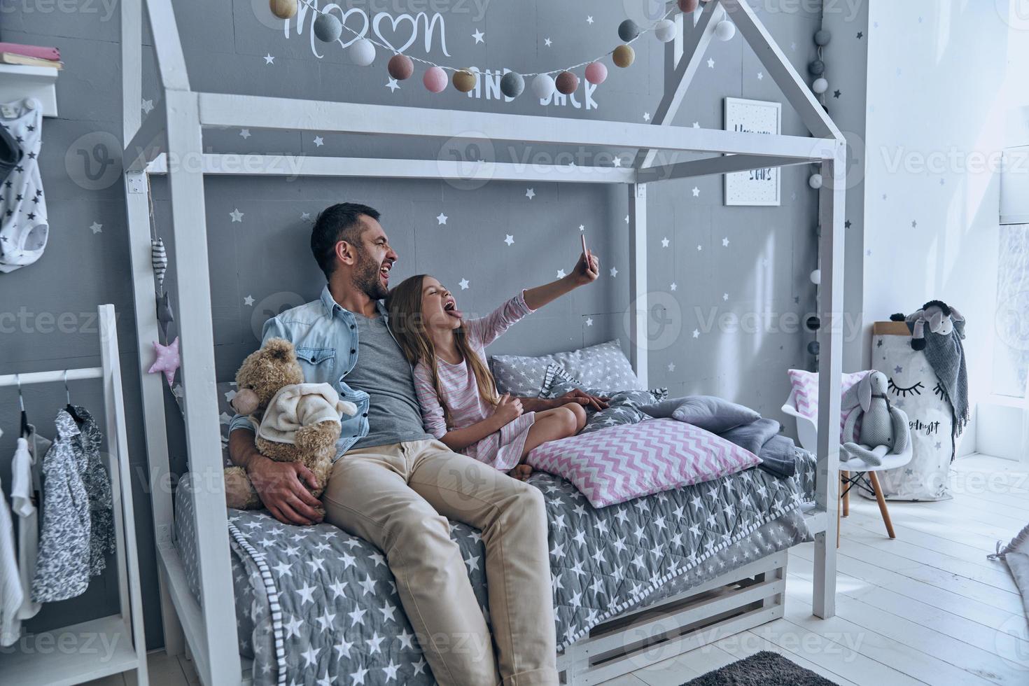 Going crazy together. Young father and his little daughter taking selfie while sitting on the bed at home photo