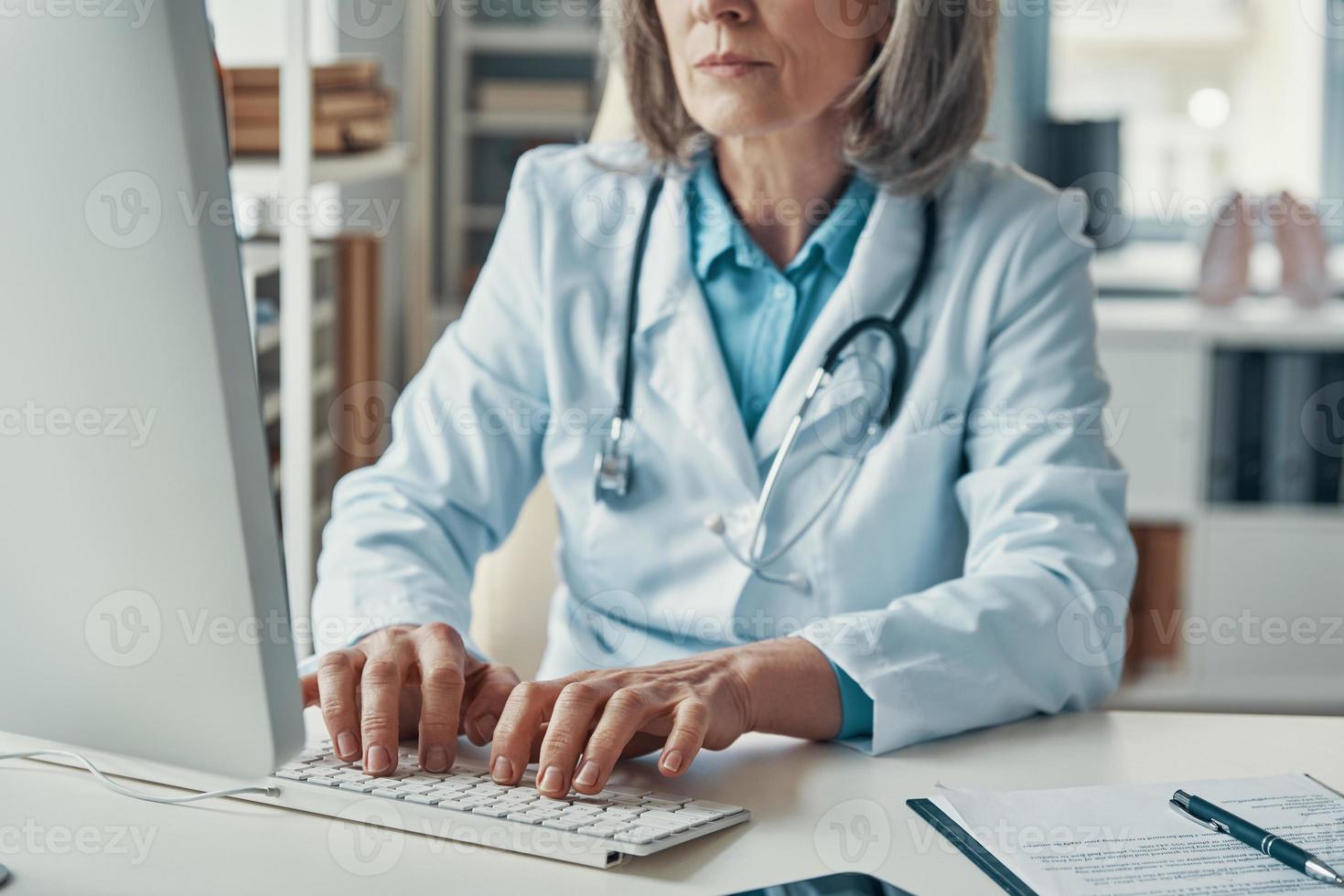 Close up of female doctor in white lab coat working using computer while sitting in her office photo