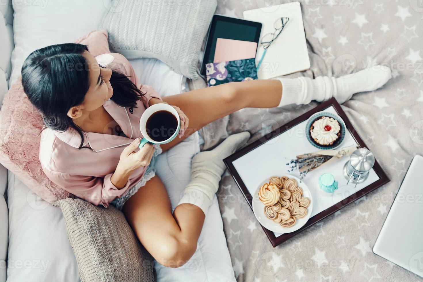 Top view of beautiful young woman in pajamas enjoying hot drink while resting in bed at home photo