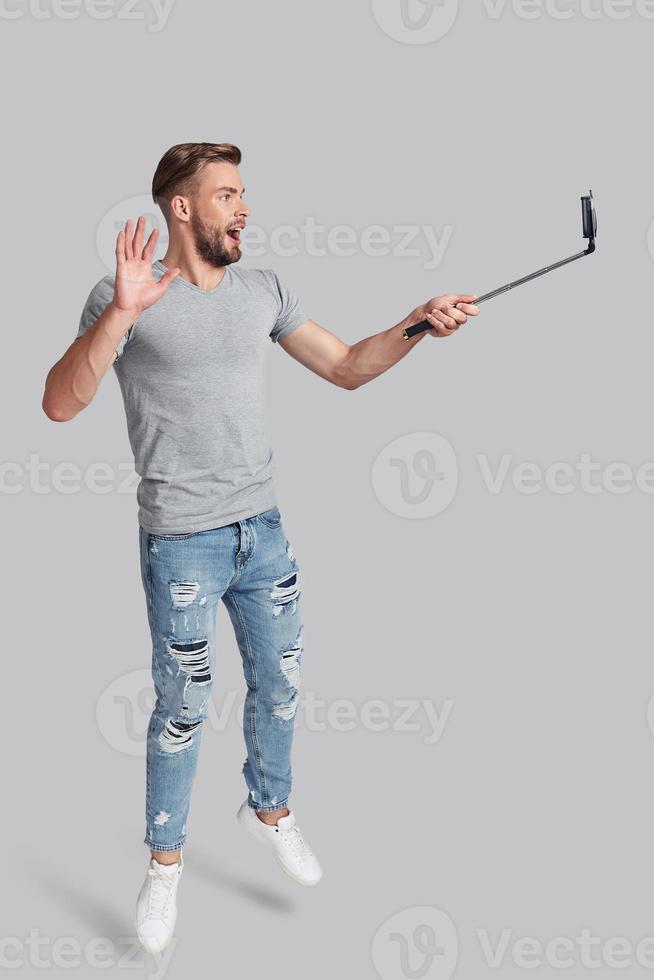 Mid-air selfie. Full length of handsome young man taking selfie while jumping against grey background photo