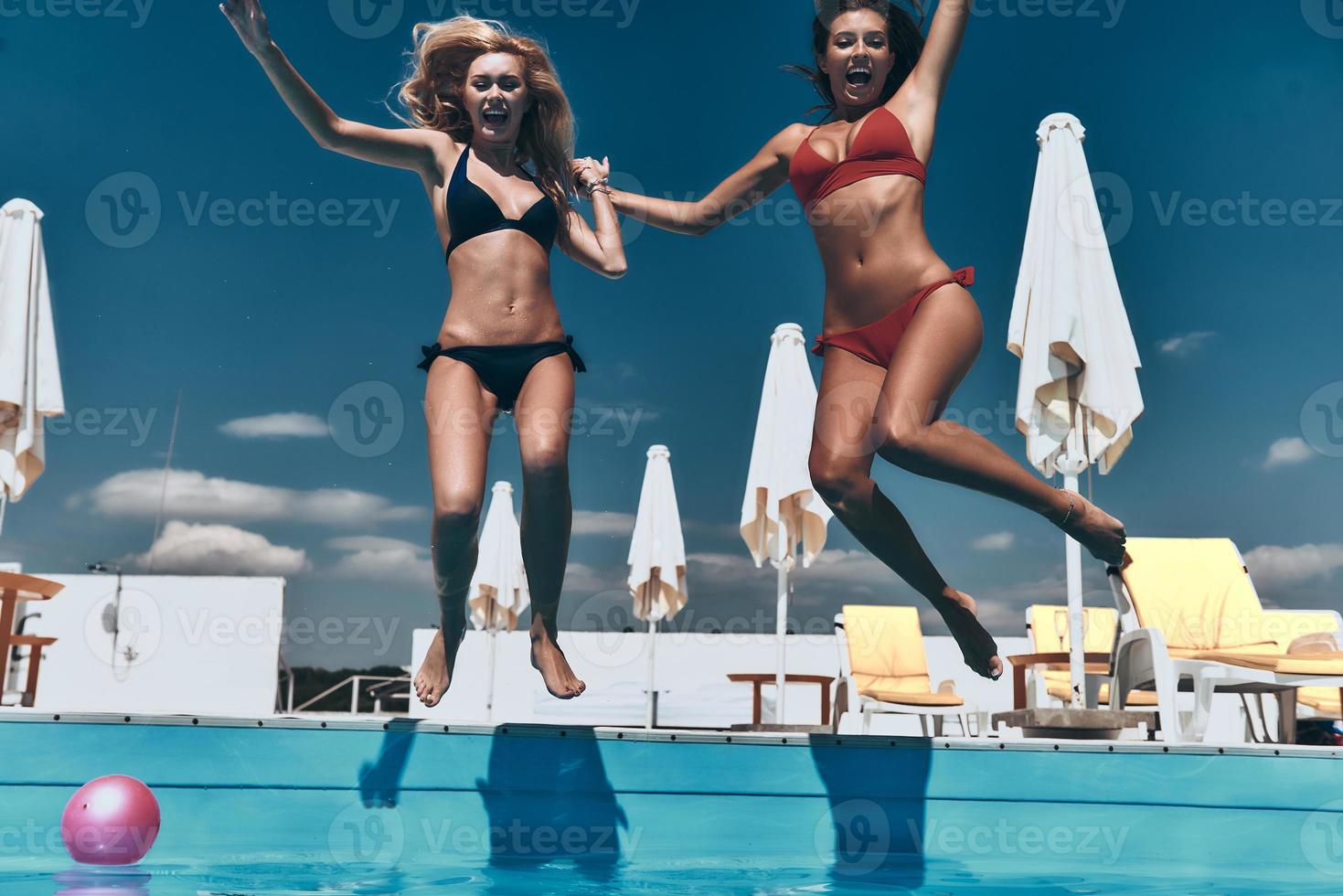 Crazy girls. Two attractive young women in bikini holding hands and keeping mouths open while jumping in the pool outdoors photo