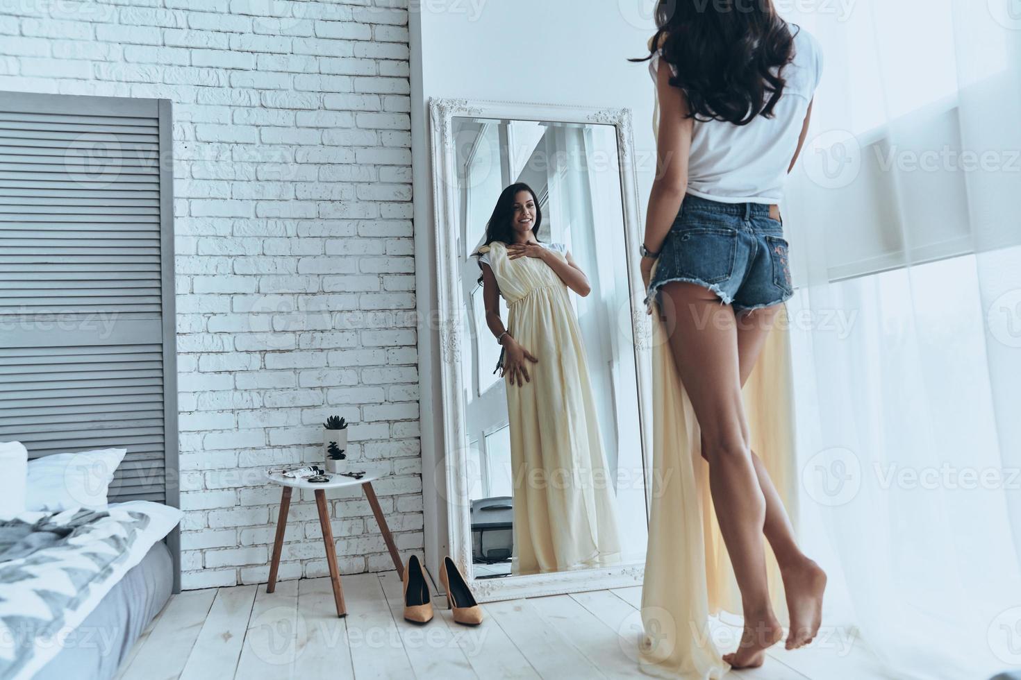 Rear view of attractive young woman trying on her dress while looking in the mirror at home photo