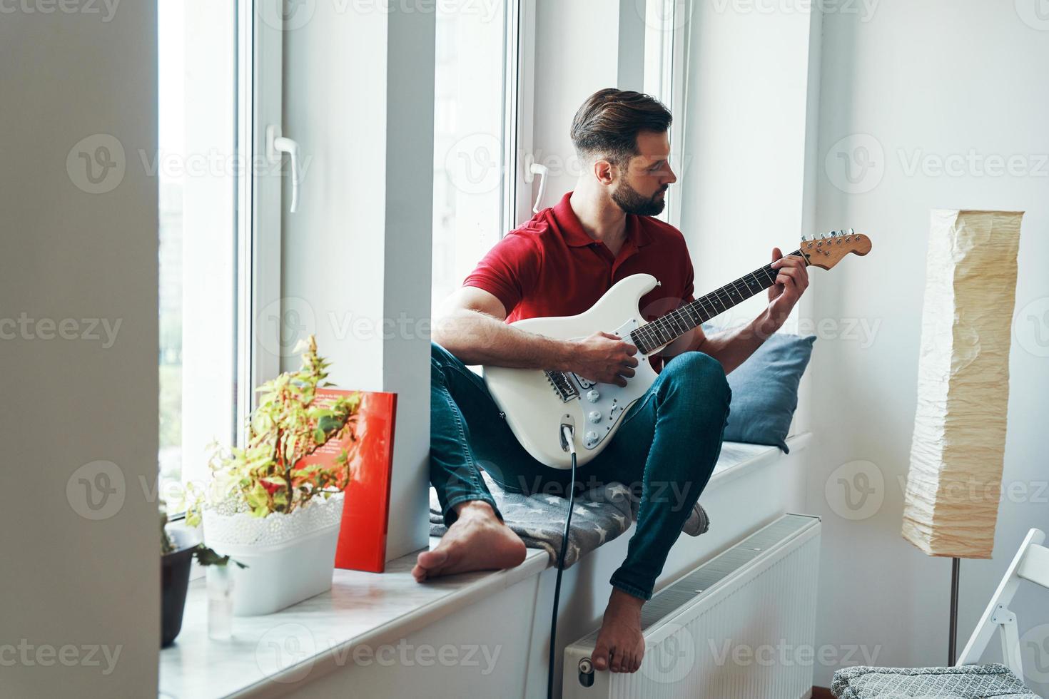 Charming young man in casual clothing playing guitar while sitting on the window sill photo