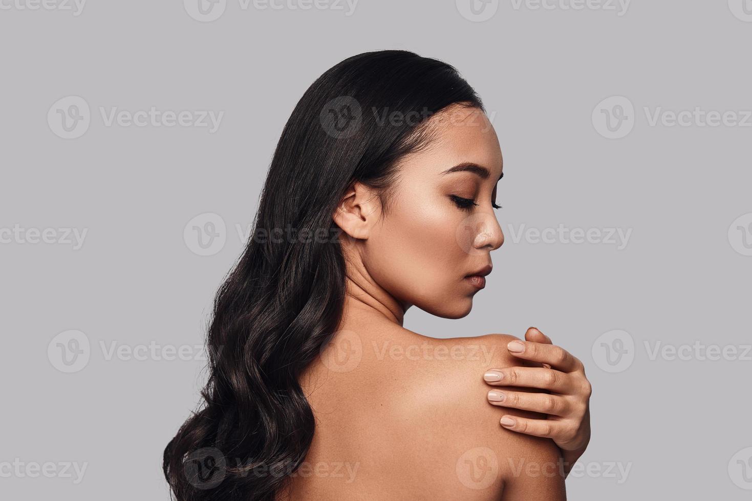 Perfect skin. Beautiful young woman touching her shoulder with hand while standing against grey background photo