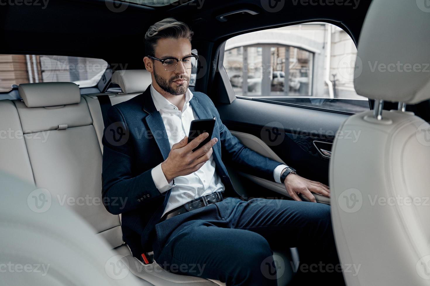 Young perfectionist. Handsome young man in full suit looking at his smart phone while sitting in the car photo
