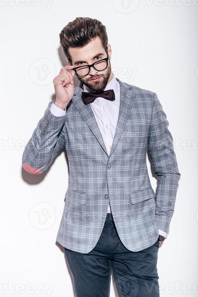 How you doing Handsome young man wearing suit keeping hand in pocket and looking over his glasses while standing against white background photo