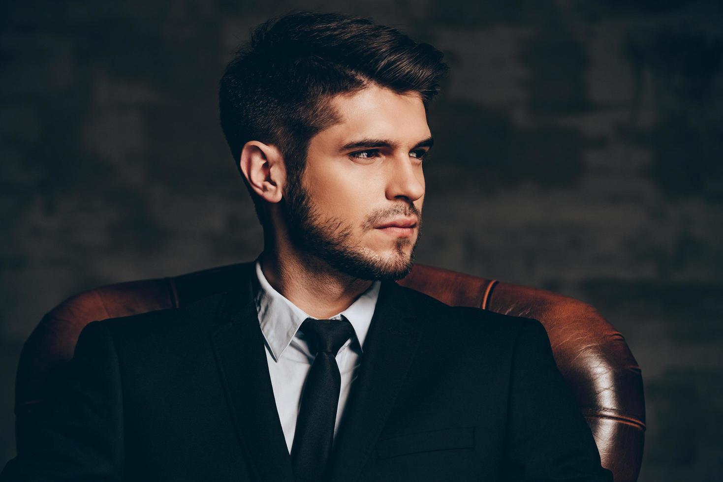 Modern man.Portrait of young handsome man in suit holding his sunglasses and looking at camera while sitting in leather chair against dark grey background photo