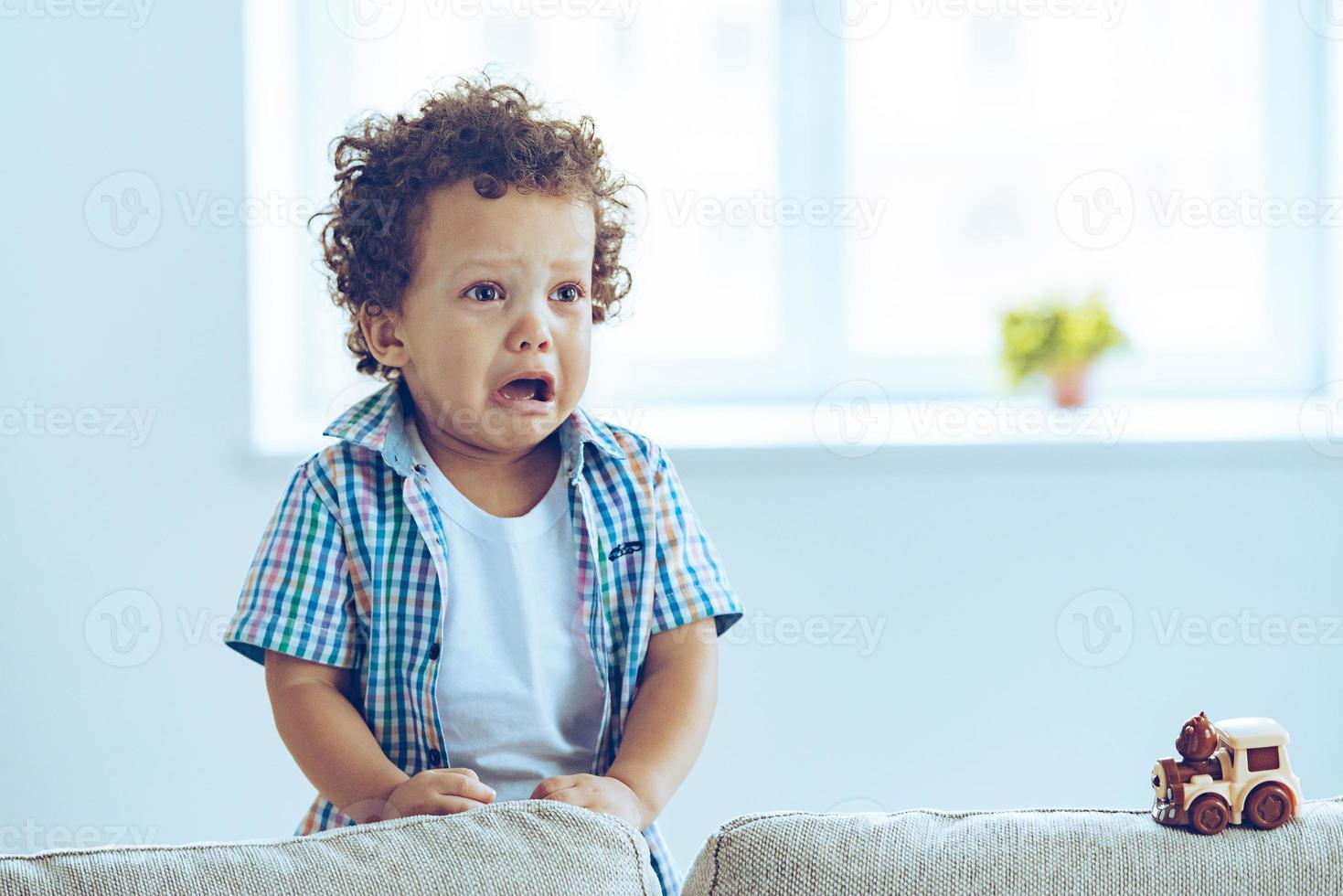 I want to play Little African baby boy crying and looking away while standing on the couch at home photo