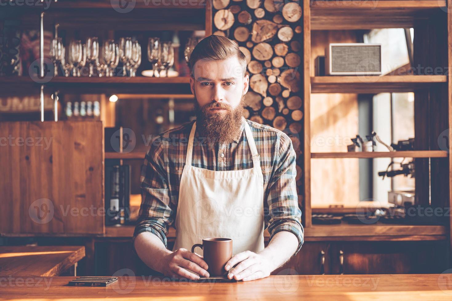 Confident barista. Young bearded man in apron looking at camera and holding coffee cup while standing at bar counter photo