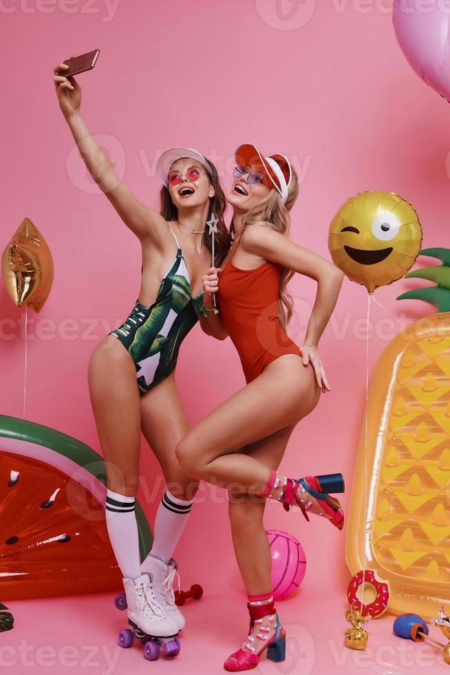 This one goes in frame. Full length of two beautiful young women in swimwear taking selfie and smiling while standing against pink background photo