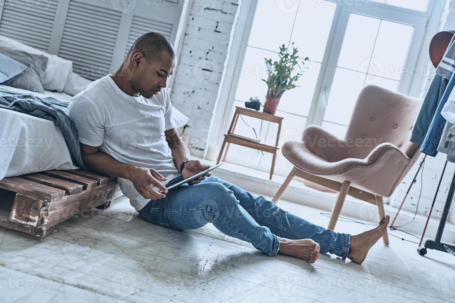 Searching for fresh news. Handsome young African man using digital tablet while flooring in the bedroom at home photo