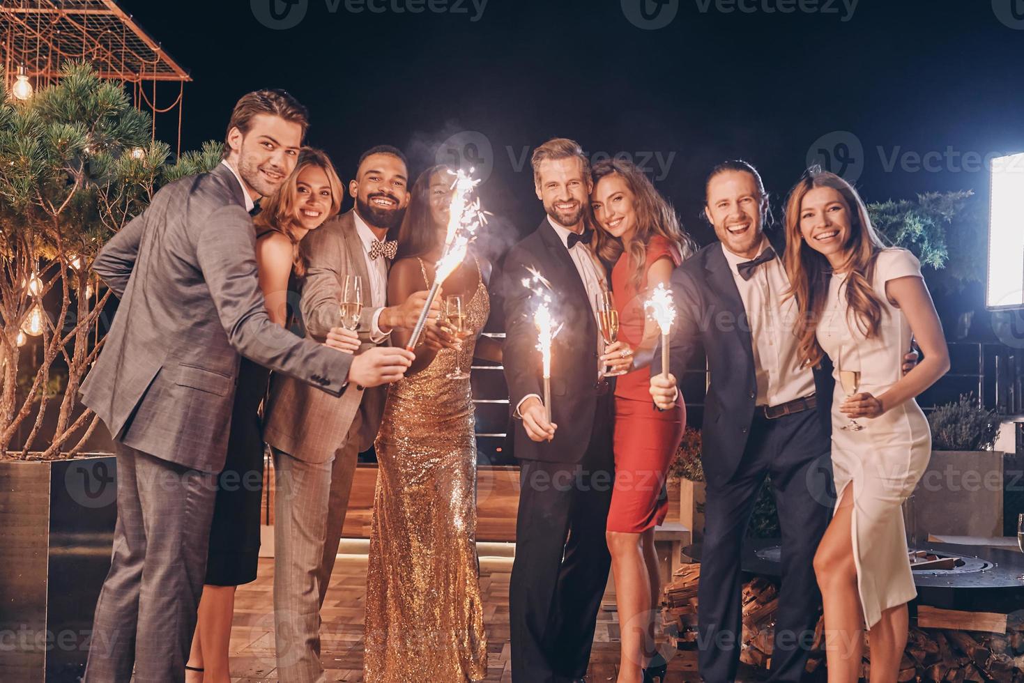 Group of beautiful people in formalwear holding sparklers and smiling while spending time on outdoor party photo