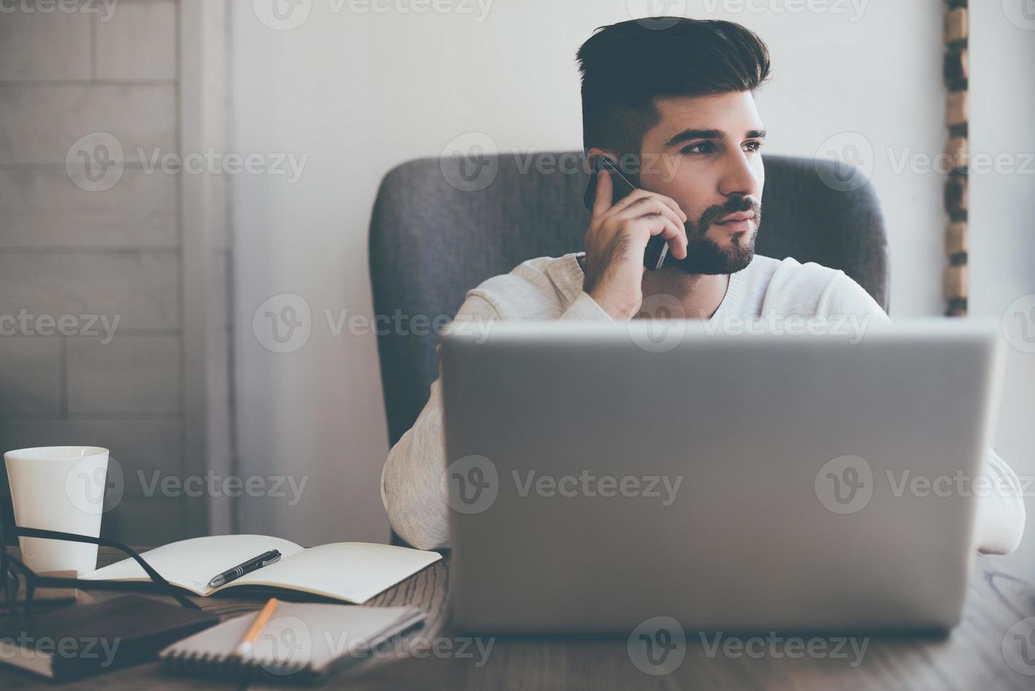 Staying in touch. Front view of thoughtful young man talking on mobile phone while sitting at his working place in office photo