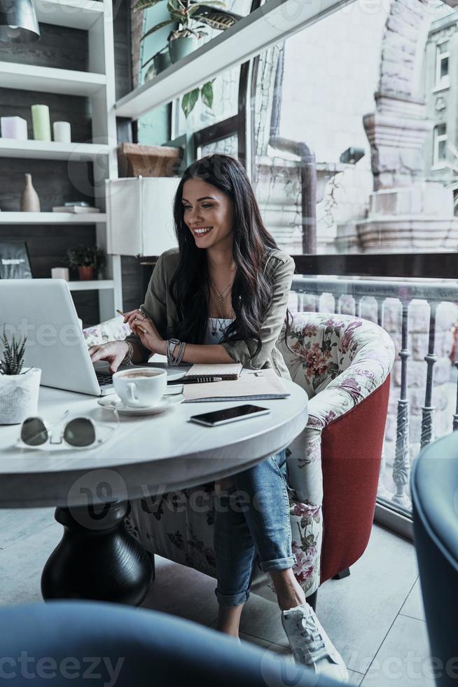 Her job is her hobby. Beautiful young woman using laptop and smiling while sitting in restaurant photo