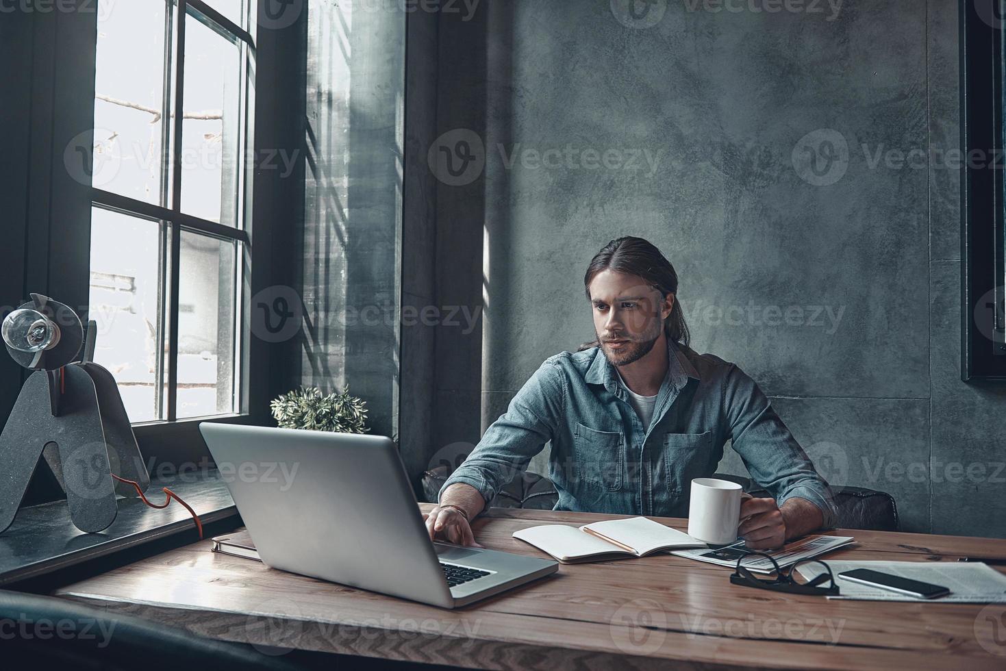 Taking care of business. Thoughtful young man working using computer and holding a cup while sitting at the office desk photo
