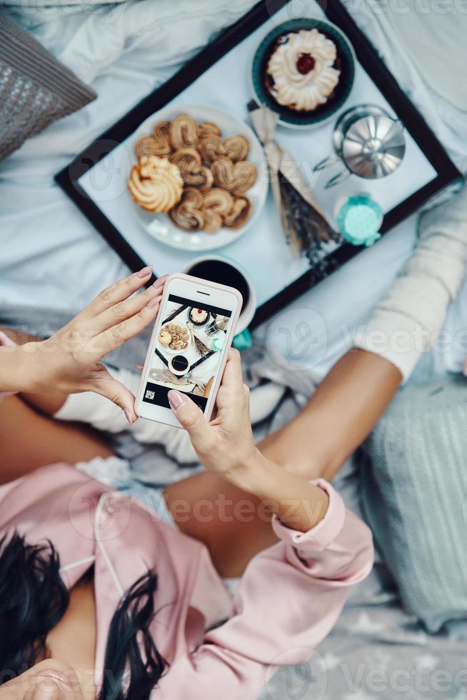 Close up top view of young woman in pajamas photographing flat lay of breakfast while resting in bed at home photo
