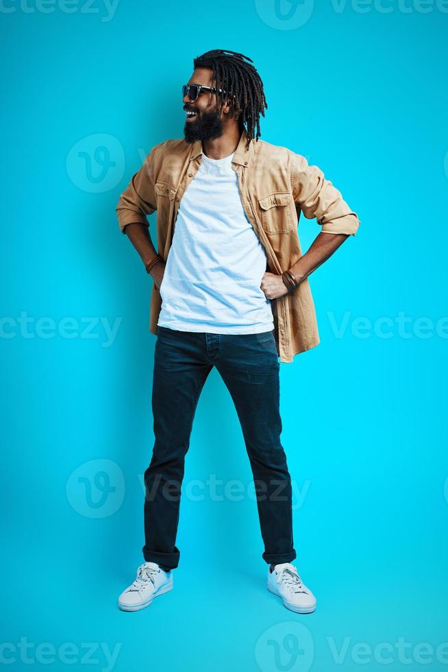 Full length of handsome young African man in casual wear looking away while standing against blue background photo