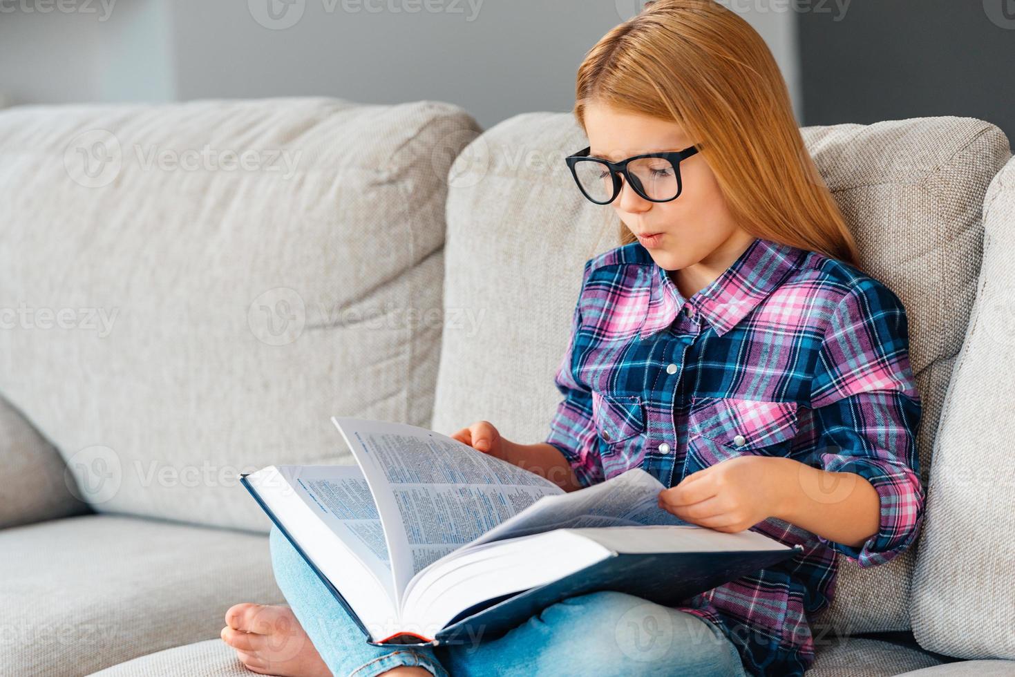 Where was that chapter Beautiful little girl in glasses reading book while sitting on the couch in lotus position at home photo