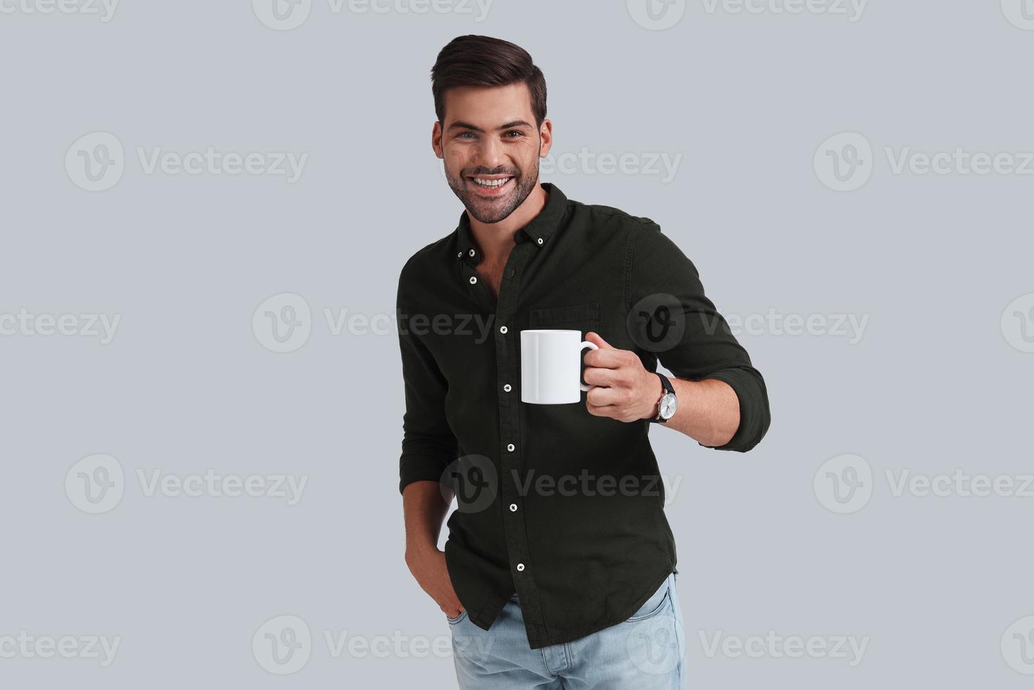 Enjoying fresh coffee.  Good looking young man in smart casual wear holding a cup and smiling while standing against grey background photo