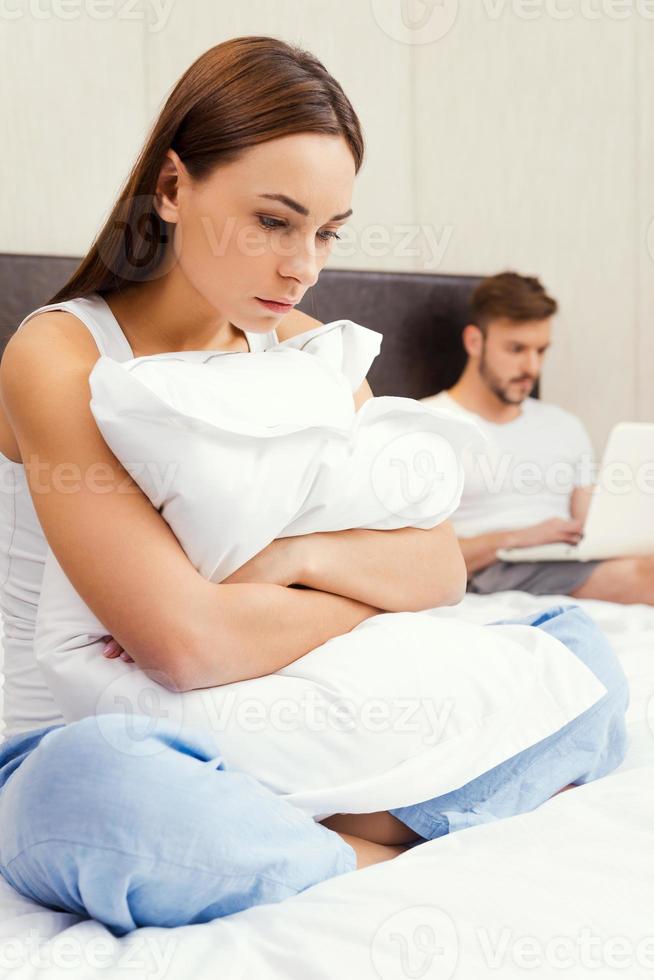 Feeling lonely near him. Depressed young woman embracing pillow and looking down while sitting on bed with young man working on laptop in the background photo